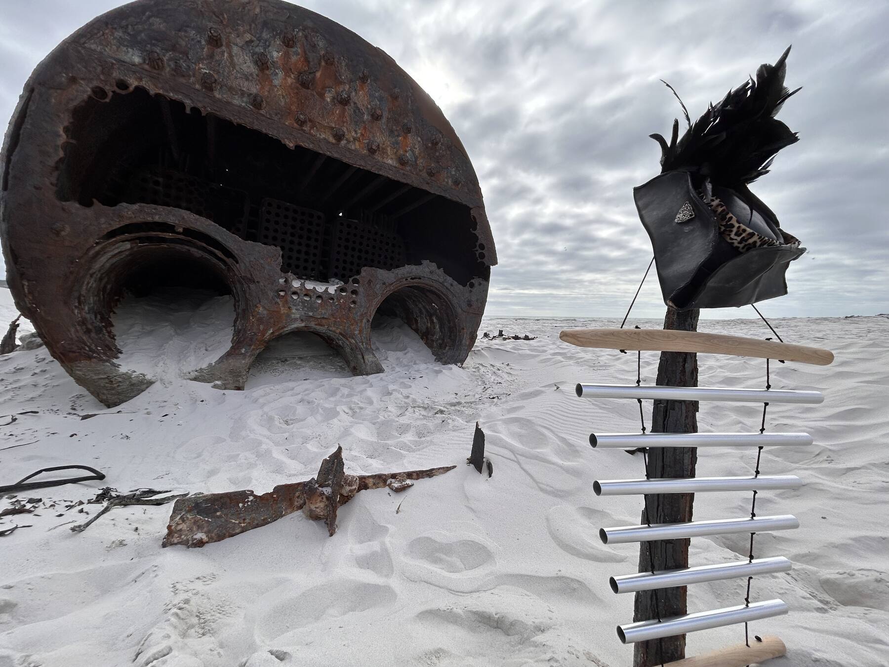 Ein rostiger Schiffskessel an einem weißen Strand, daneben ein Koshi-Klangspiel an einem Pfahl, darauf ein Dreispitz aus schwarzen Federn