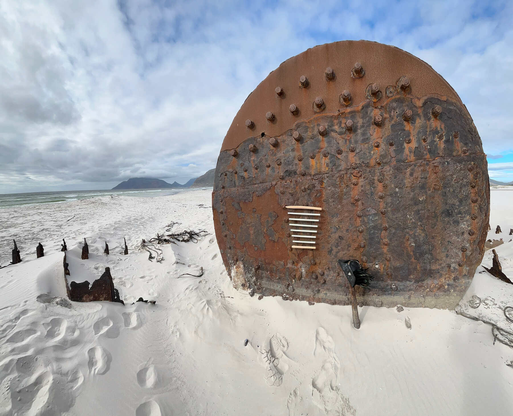 Ein rostiger Schiffskessel an einem weissen Strand, daran haengt ein Koshi-Klangspiel, davor steckt ein Dreispitz im Sand
